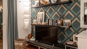 A living room with a TV, shelves with decorative items, a wicker basket, and a patterned wall.