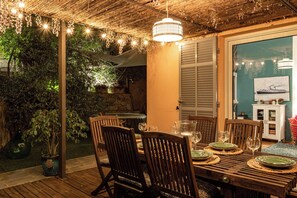 An outdoor dining area with a wooden table set for four, surrounded by chairs, under a thatched roof with hanging lights.