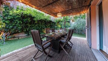 Une terrasse extérieure en bois, avec une table et des chaises, entourée d’une végétation luxuriante et surmontée d’un toit de chaume.