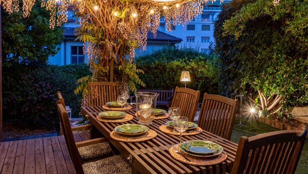 An outdoor dining area with a wooden table set for four, surrounded by chairs, under a bamboo canopy with hanging lights.