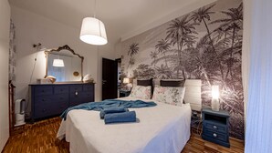 A bedroom with a bed, a dresser, a nightstand, a lamp, and a wall with a tropical mural.