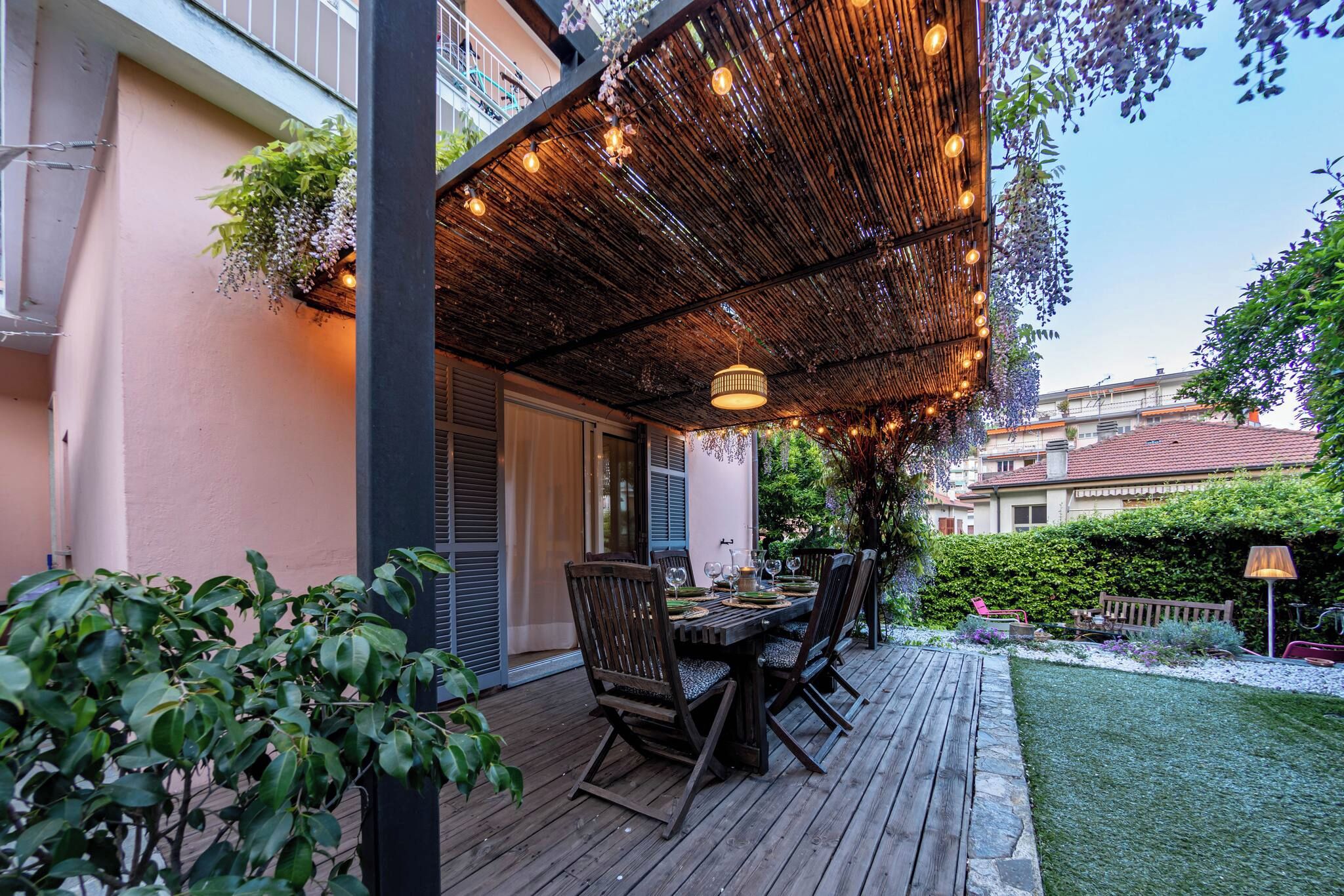 An outdoor patio with a wooden table set for dining, surrounded by chairs, under a bamboo canopy with hanging lights.