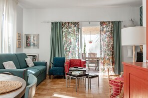A living room with a sofa, chairs, a table, and a view through a window with curtains.