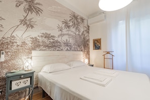 A bedroom with a bed, a nightstand, a lamp, a wall-mounted air conditioner, and a painting.