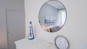 A white room with a round mirror, a clock, and a small table with a decorative anchor and leaves.