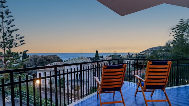 A balcony with two wooden chairs and a table, overlooking a coastal cityscape with buildings and trees.