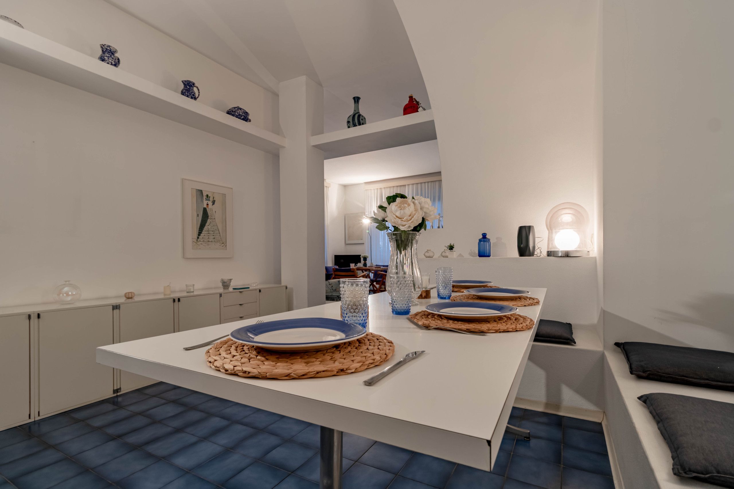 A modern dining area with a white table set for two, featuring woven placemats and blue plates.