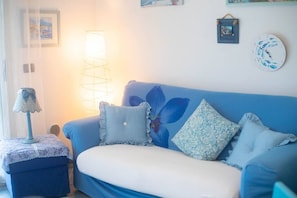 Un sofá azul con cojines estampados, un reposapiés con encaje, una lámpara de pared y un reloj.