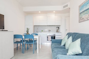 A compact living space with a kitchenette, a dining area with blue chairs, and a grey sofa with cushions.