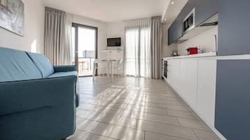 A modern living room with a sofa, a dining area with chairs, a kitchen with cabinets and appliances, and large windows with curtains.