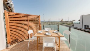 Un balcó amb una taula de fusta preparada per a dues persones, caires blanques i vista al mar amb embarcacions a la distància.