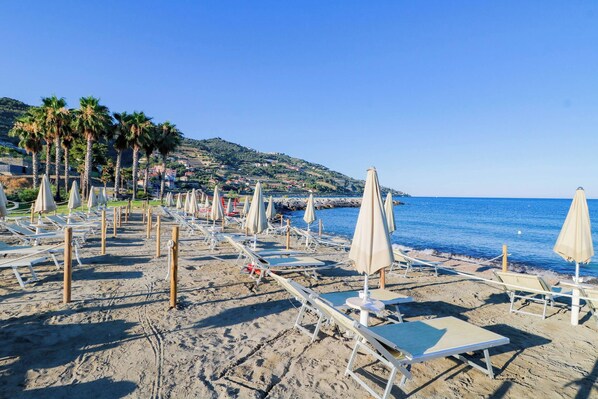 A beach with rows of sun loungers and umbrellas, a clear blue sky, and a hilly landscape in the background.