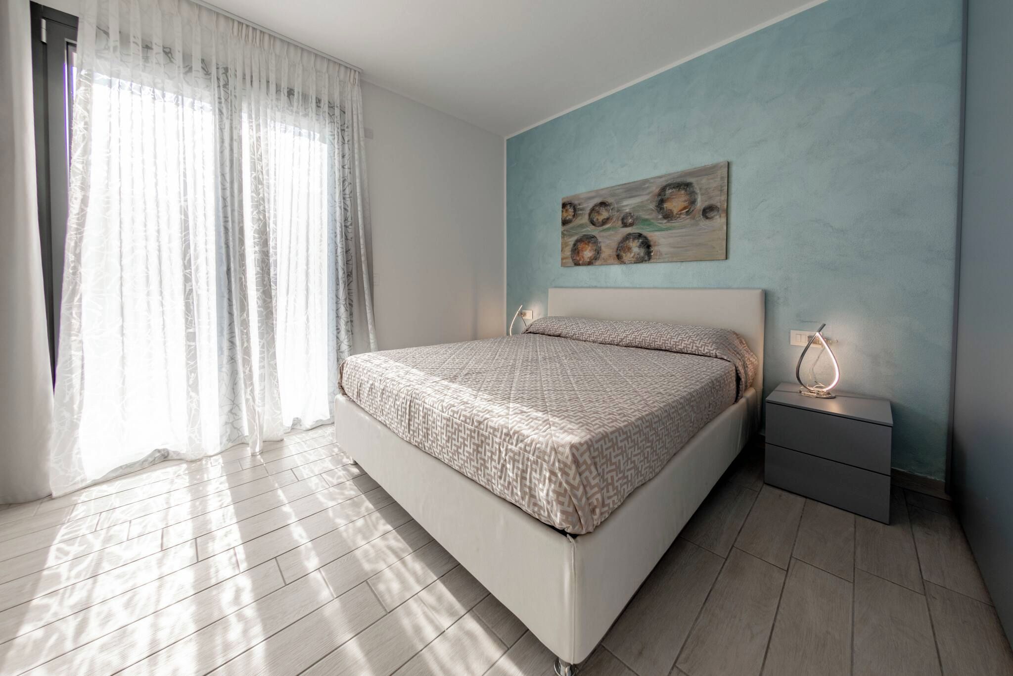 Un dormitorio moderno con una cama grande, una mesita de noche con lámpara y un ventanal con cortinas traslúcidas.
