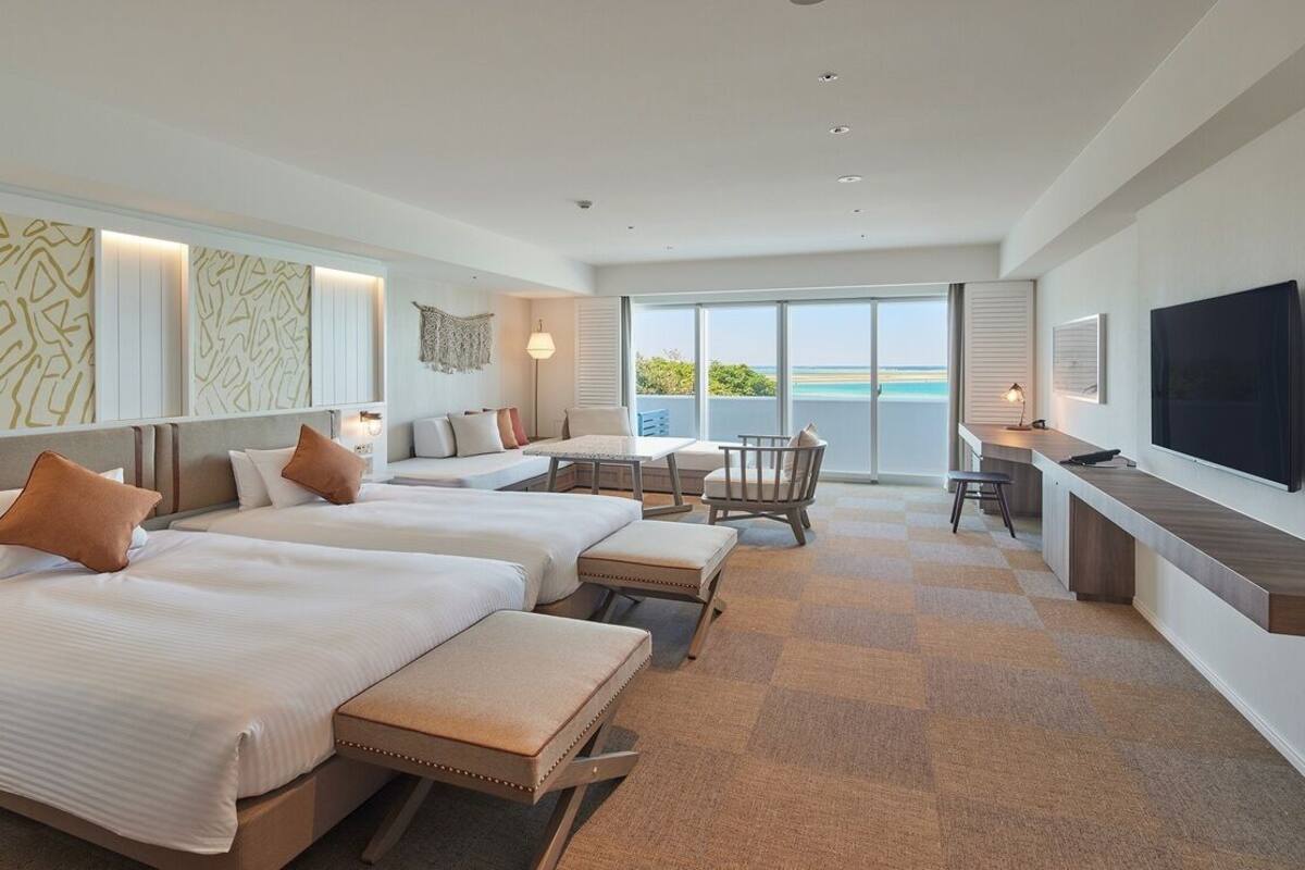 Habitación de hotel con dos camas, zona de comedor, televisión y balcón con vistas.