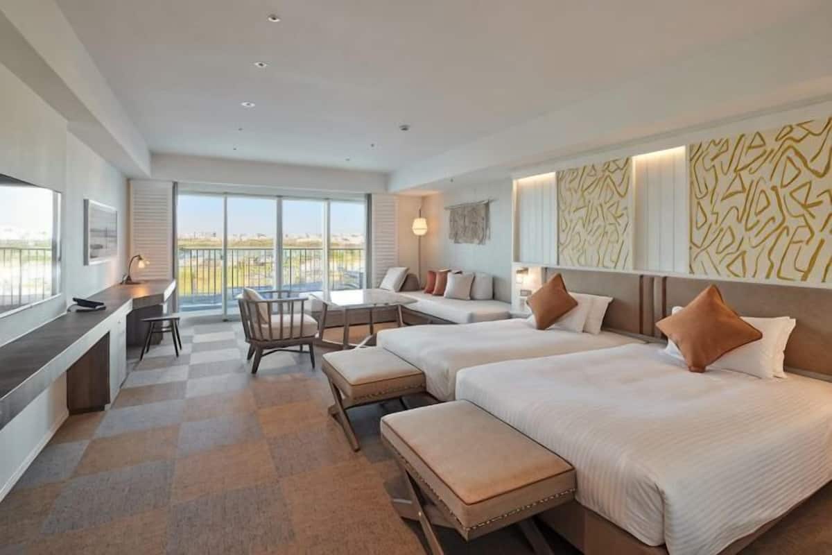 A hotel room with two beds, a desk, a chair, a small table, and a balcony with a view.