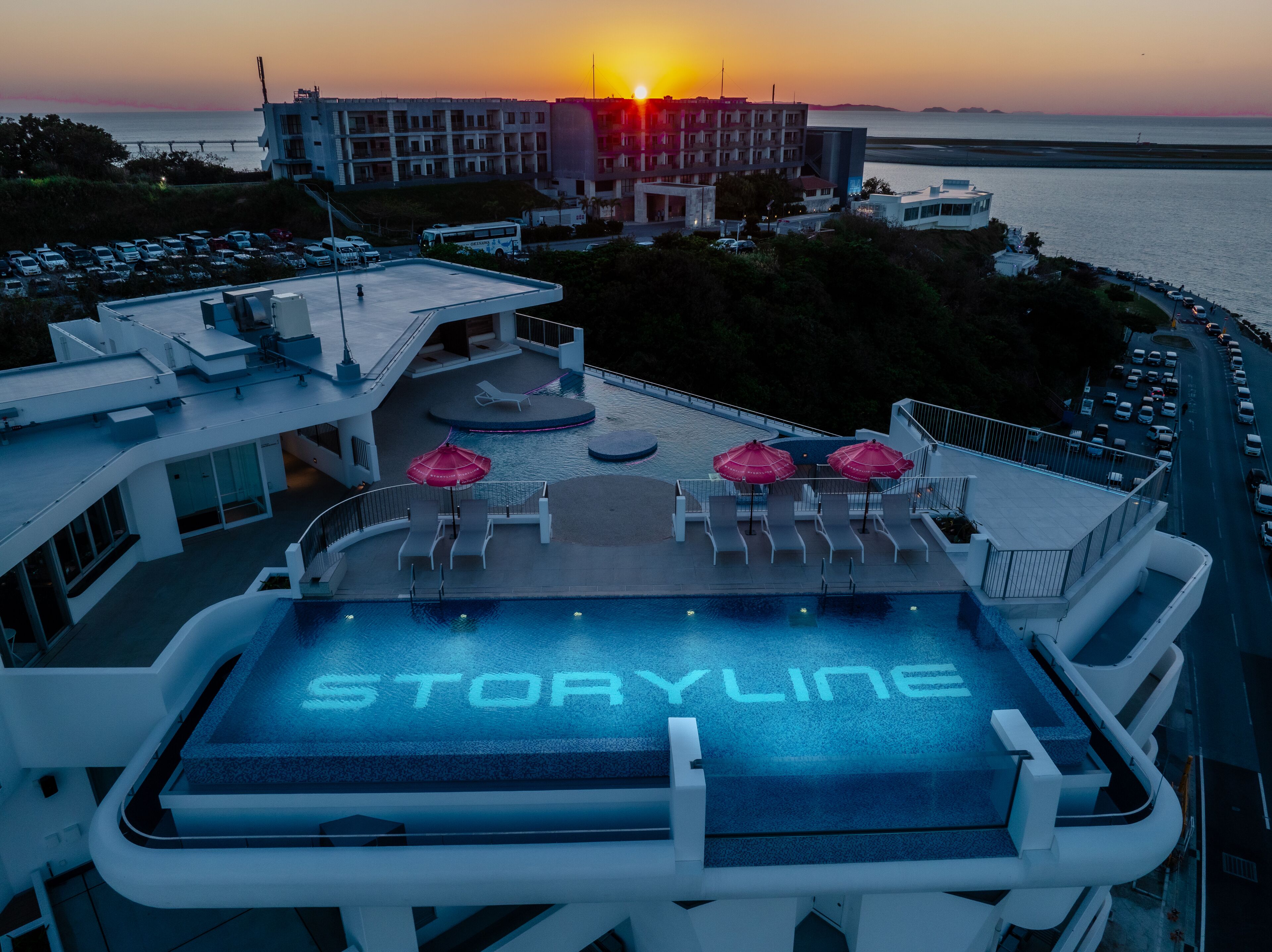 Un espace piscine aménagé sur le toit, avec le mot « STORYLINE » inscrit sur le sol, des chaises longues, des parasols et une vue sur le coucher de soleil sur l’océan.