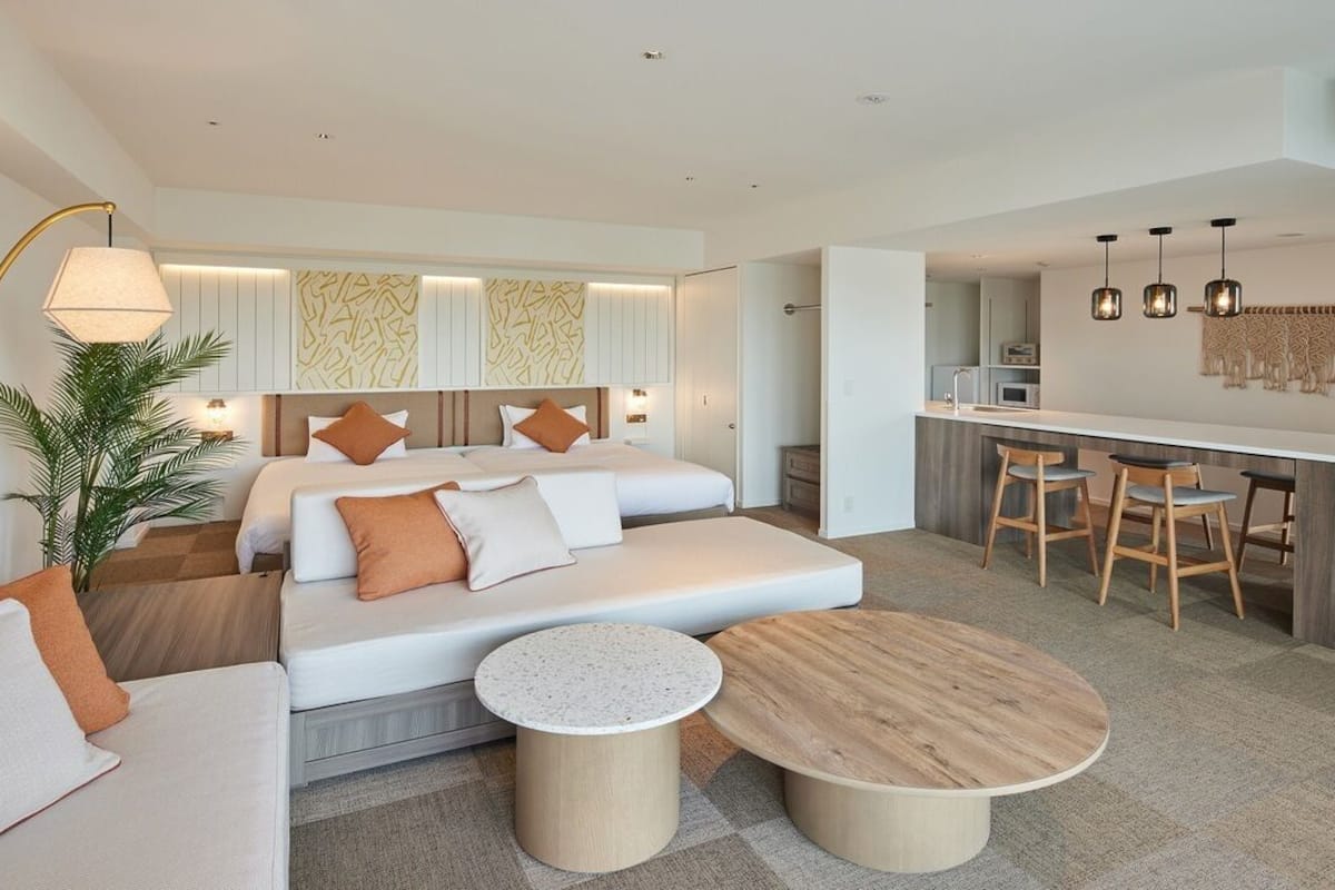 Una habitación de hotel moderna con sofá cama, zona de comedor con taburetes de bar, mesa redonda de madera y un ventanal grande.