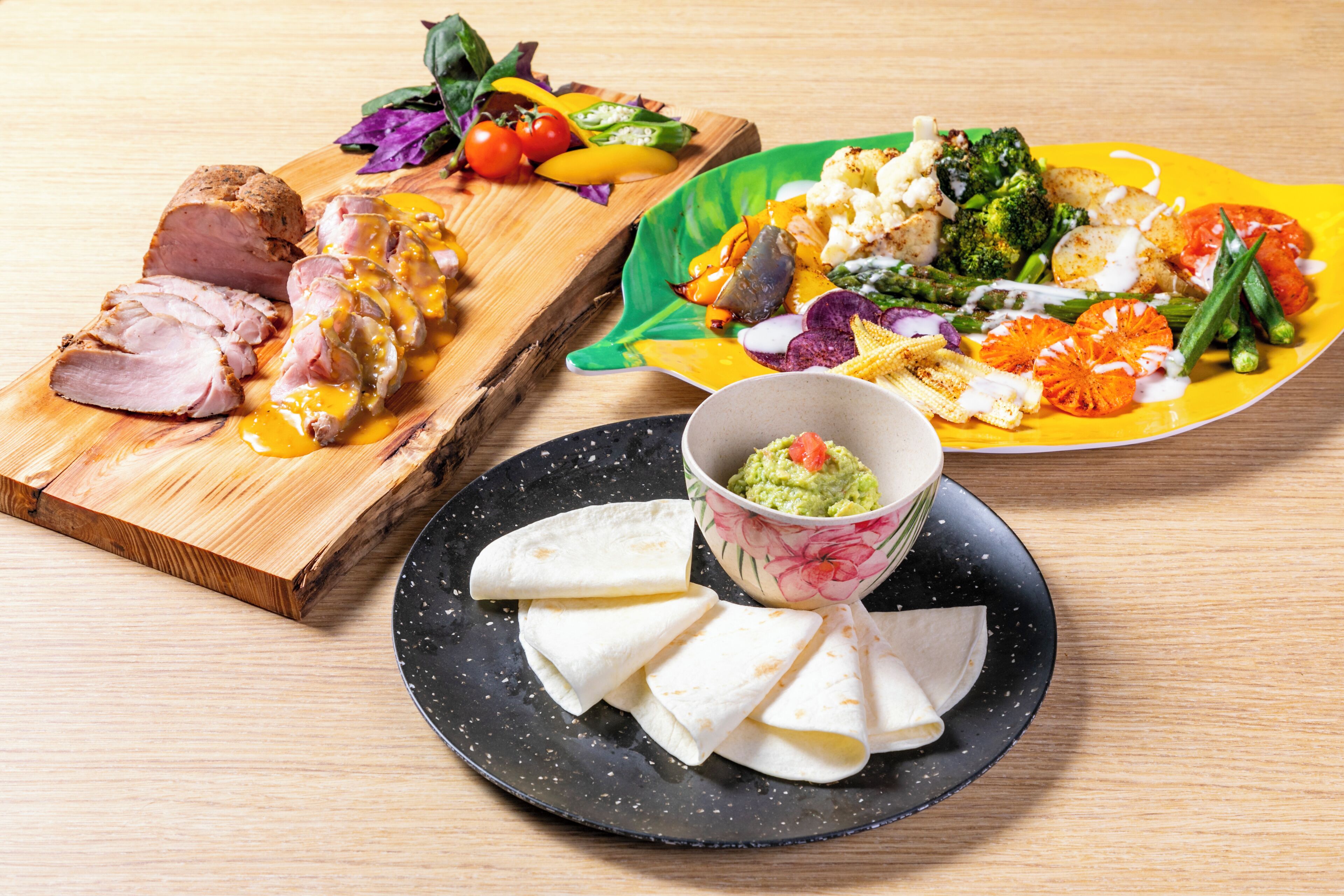 A wooden cutting board with sliced meat and vegetables, a colorful plate with a variety of vegetables and a bowl of guacamole, and a black plate with tortillas and a bowl of guacamole.