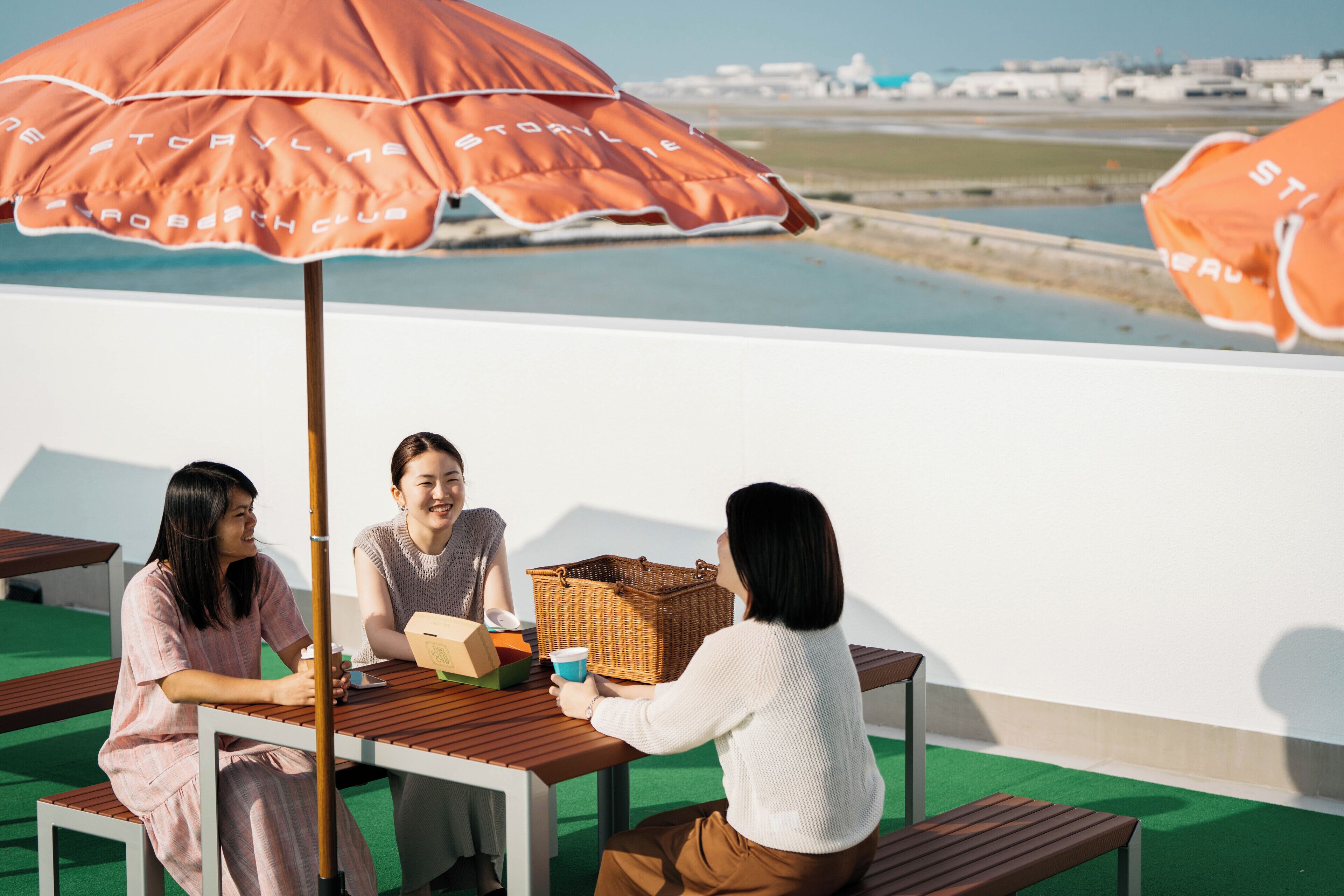 Three people seated at a wooden table with a wicker basket and a cup, under an orange umbrella with white text.