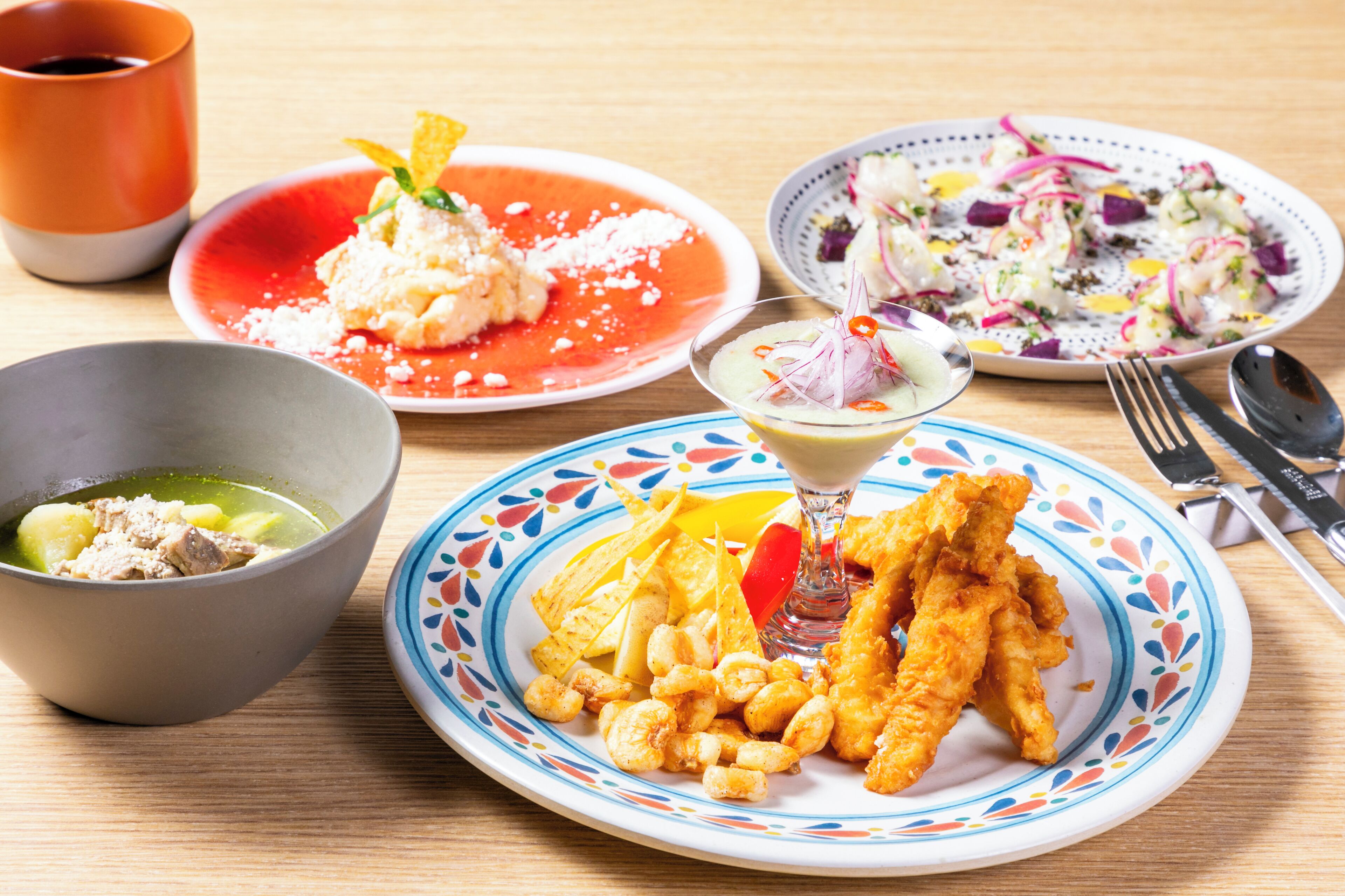 A table with various dishes including a bowl of soup, a plate of fried fish, a plate of ceviche, a plate of salad, and a glass of cocktail.