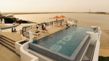 A large, rectangular infinity pool with a clear view of the sea, surrounded by white poolside furniture and orange umbrellas.