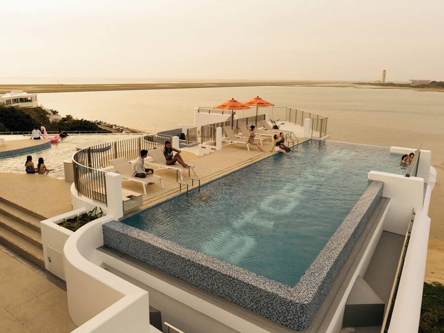 A large, rectangular infinity pool with a clear view of the sea, surrounded by white poolside furniture and orange umbrellas.