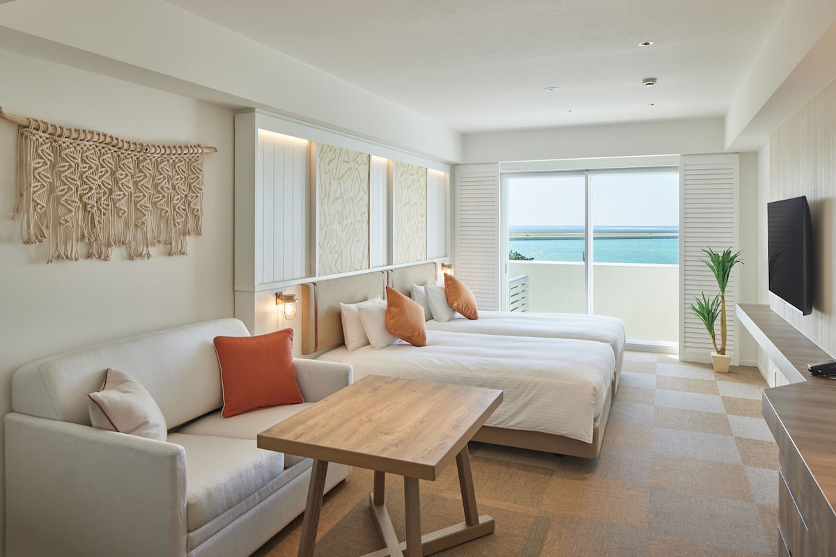 Habitación de hotel moderna con una cama grande, un sofá, una mesa de madera y un televisor. La habitación tiene un ventanal con vistas al océano.