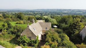 Une maison en pierre, dotée d’un toit à pente, entourée d’une végétation luxuriante et offrant une vue sur des collines ondoyantes au loin.