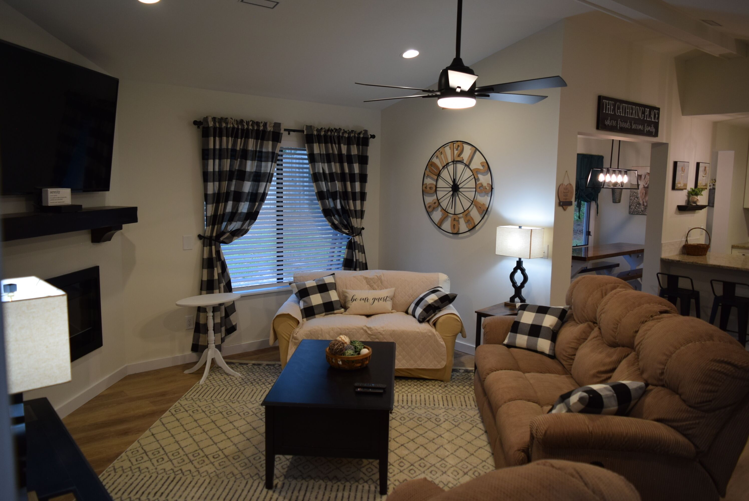 A living room with a beige sofa, a brown sofa, a black coffee table, a TV mounted on the wall, a ceiling fan, and a wall clock.