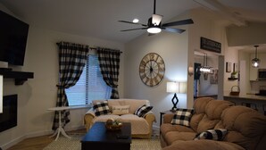 A living room with a beige sofa, a brown sofa, a coffee table, and a ceiling fan. There is a wall clock and a sign that reads "The Gathering Place".