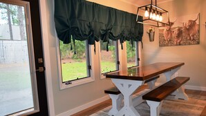 A dining area with a wooden table and benches, large windows with green valance, a chandelier, and a painting of yaks on the wall.