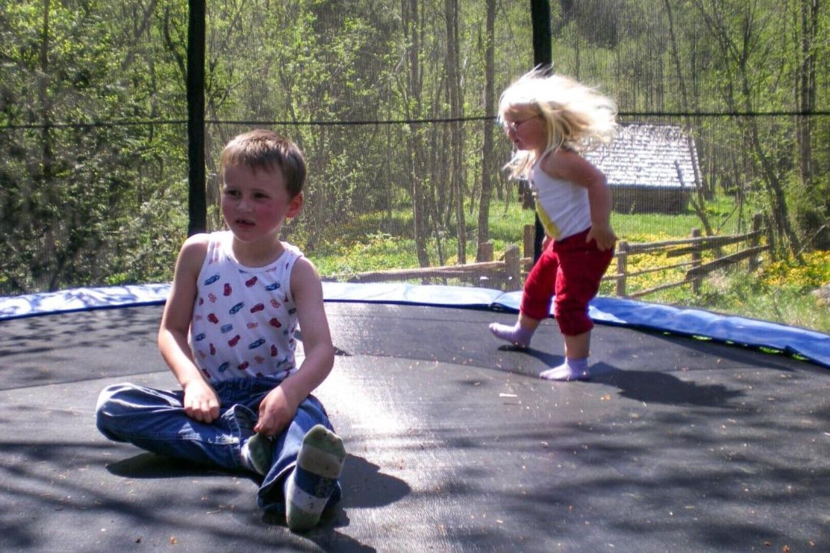 Two children on a trampoline in a fenced outdoor area with trees and a wooden fence in the background.