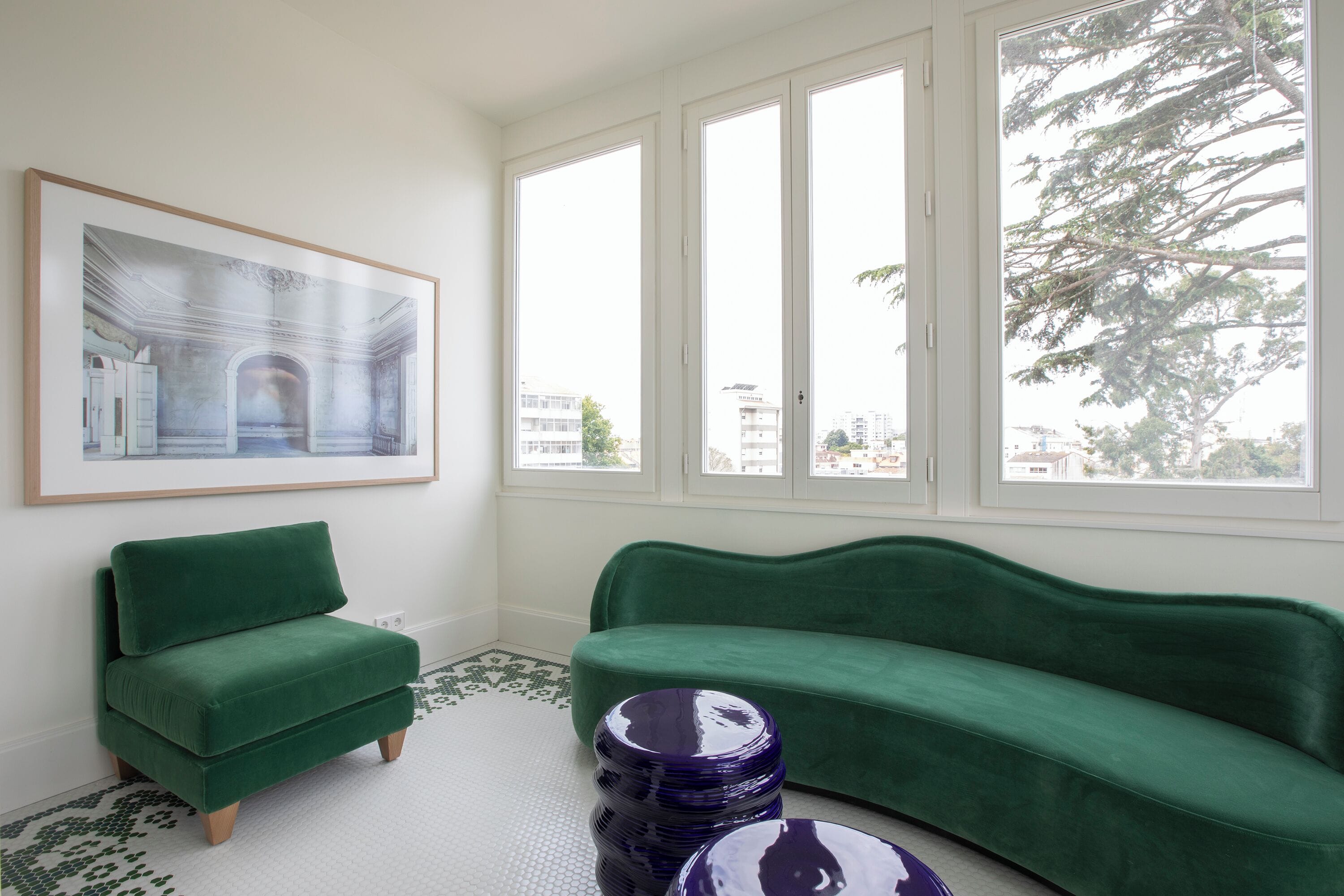 Una habitación con un sofá verde, un cuadro en la pared y ventanas amplias con vistas a edificios y árboles.