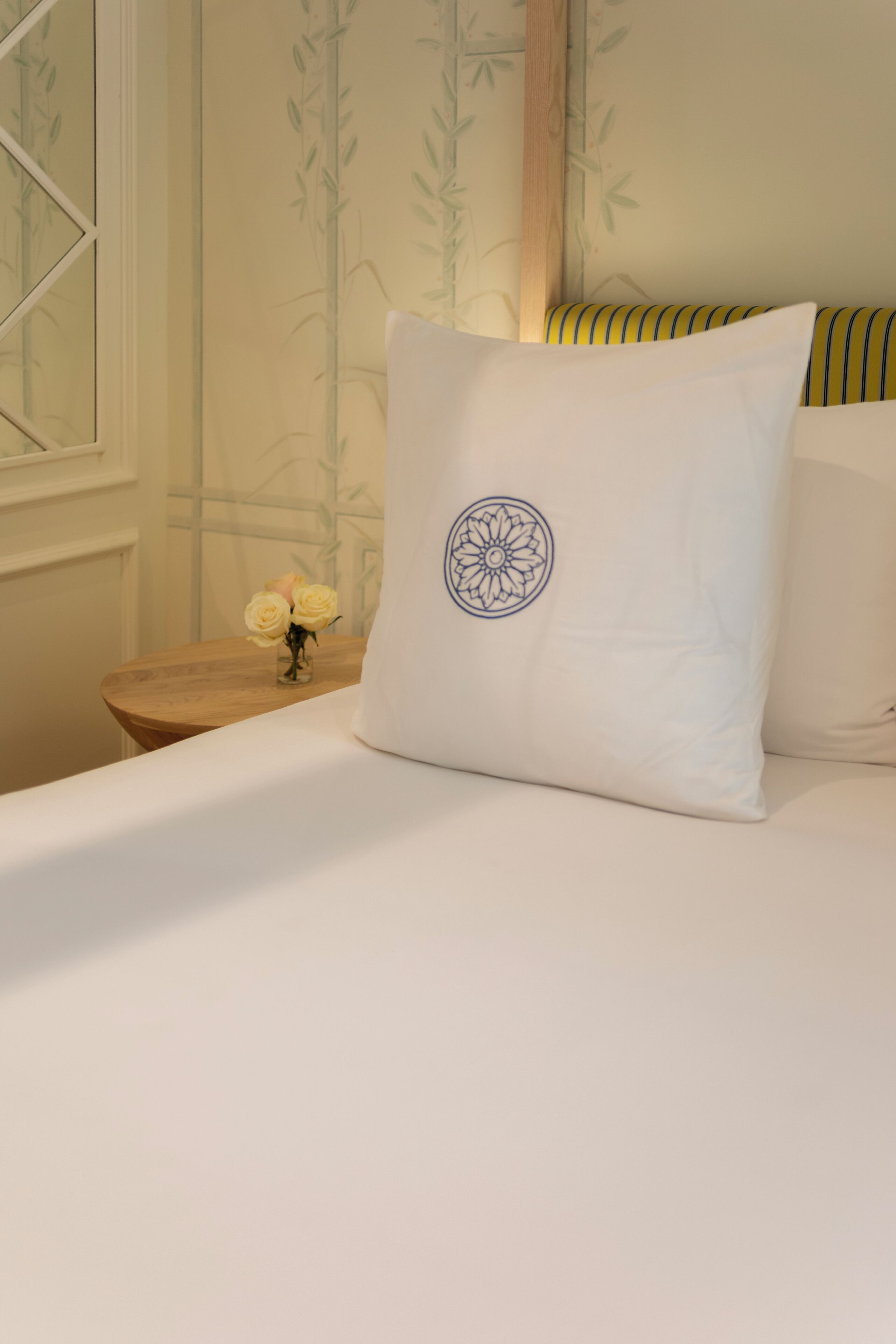 Una cama bien hecha con un almohadón blanco y un diseño de mandala azul, una mesita de noche de madera con un jarrón de rosas amarillas, y una pared con papel tapiz de bambú.