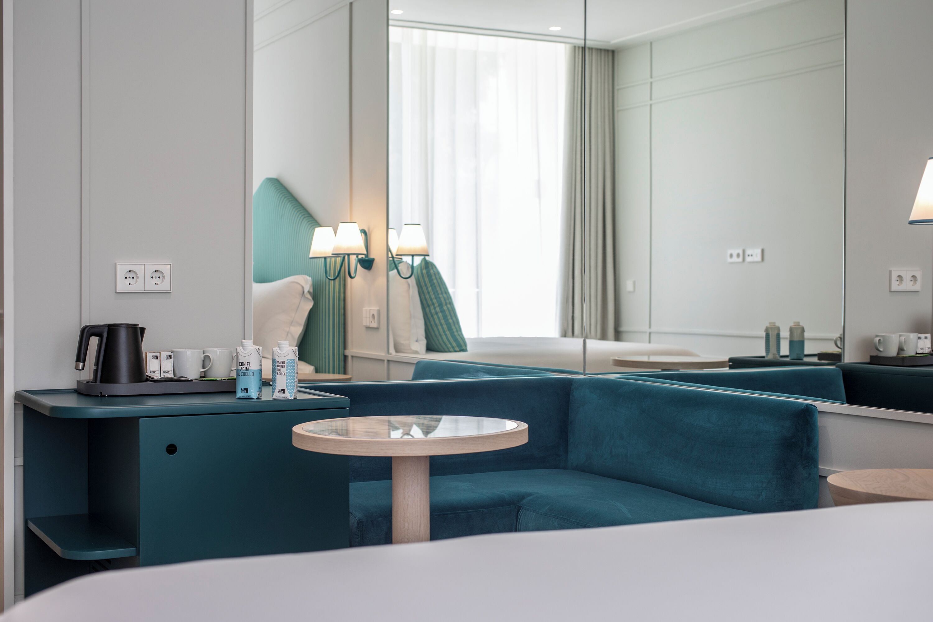A modern hotel room with a teal sofa, a round glass table, and a small bedside table with a lamp.