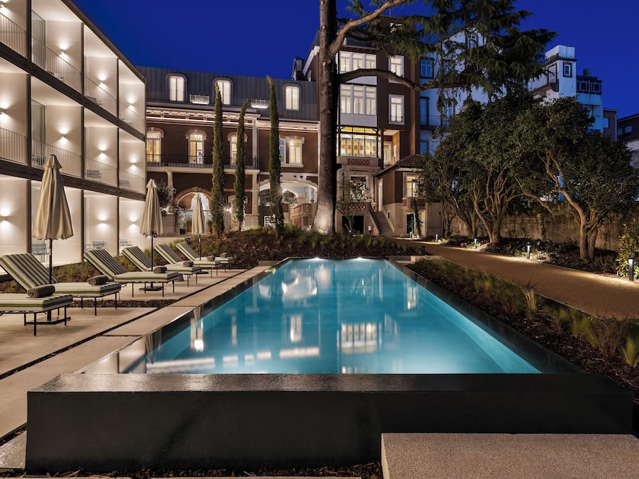 Zona de piscina nocturna con sillones de descanso y sombrillas, rodeada de edificios de varios pisos con ventanas iluminadas.