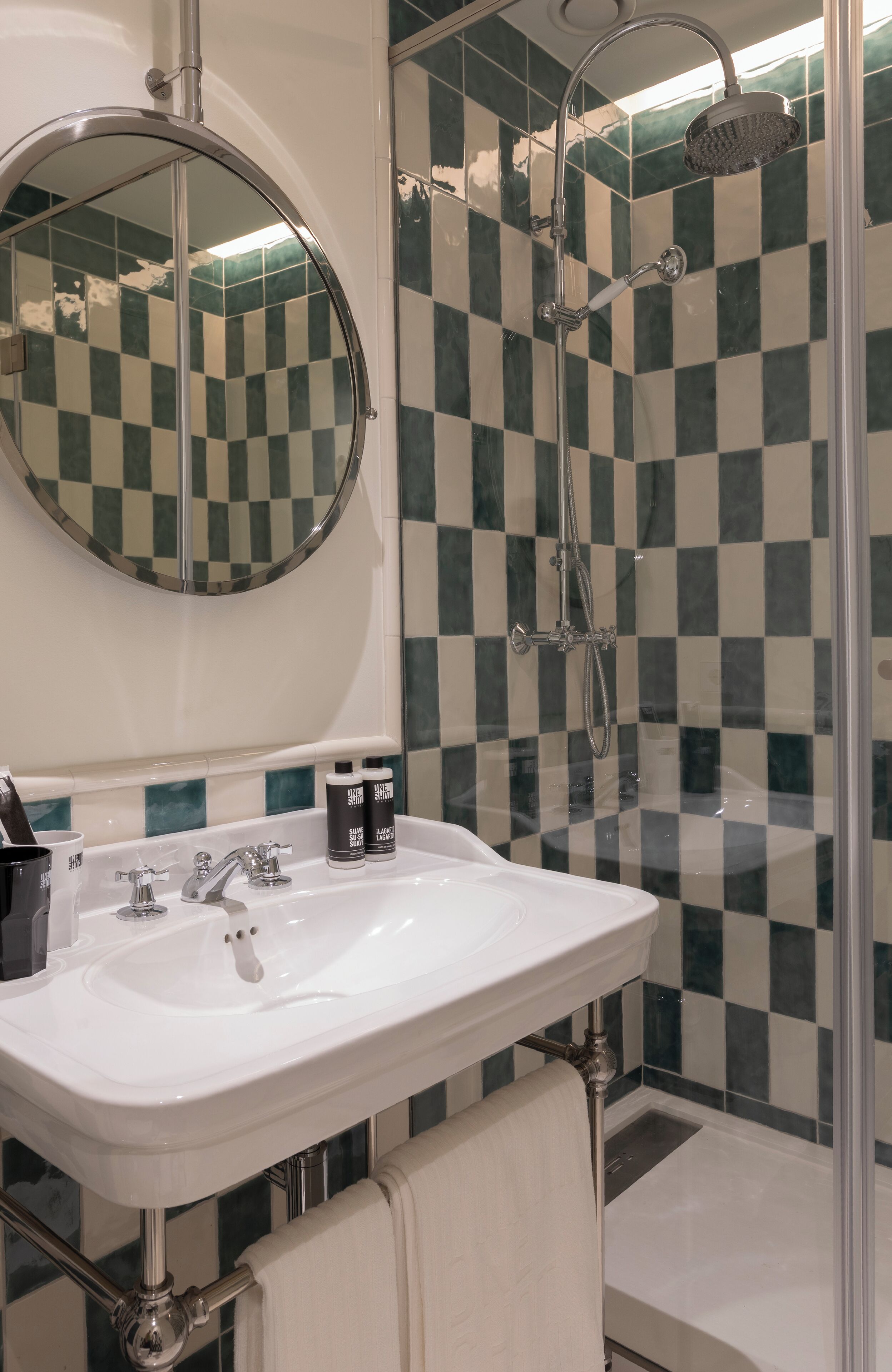 A bathroom with a checkered tile wall, a round mirror, a sink with a faucet, and a shower area.