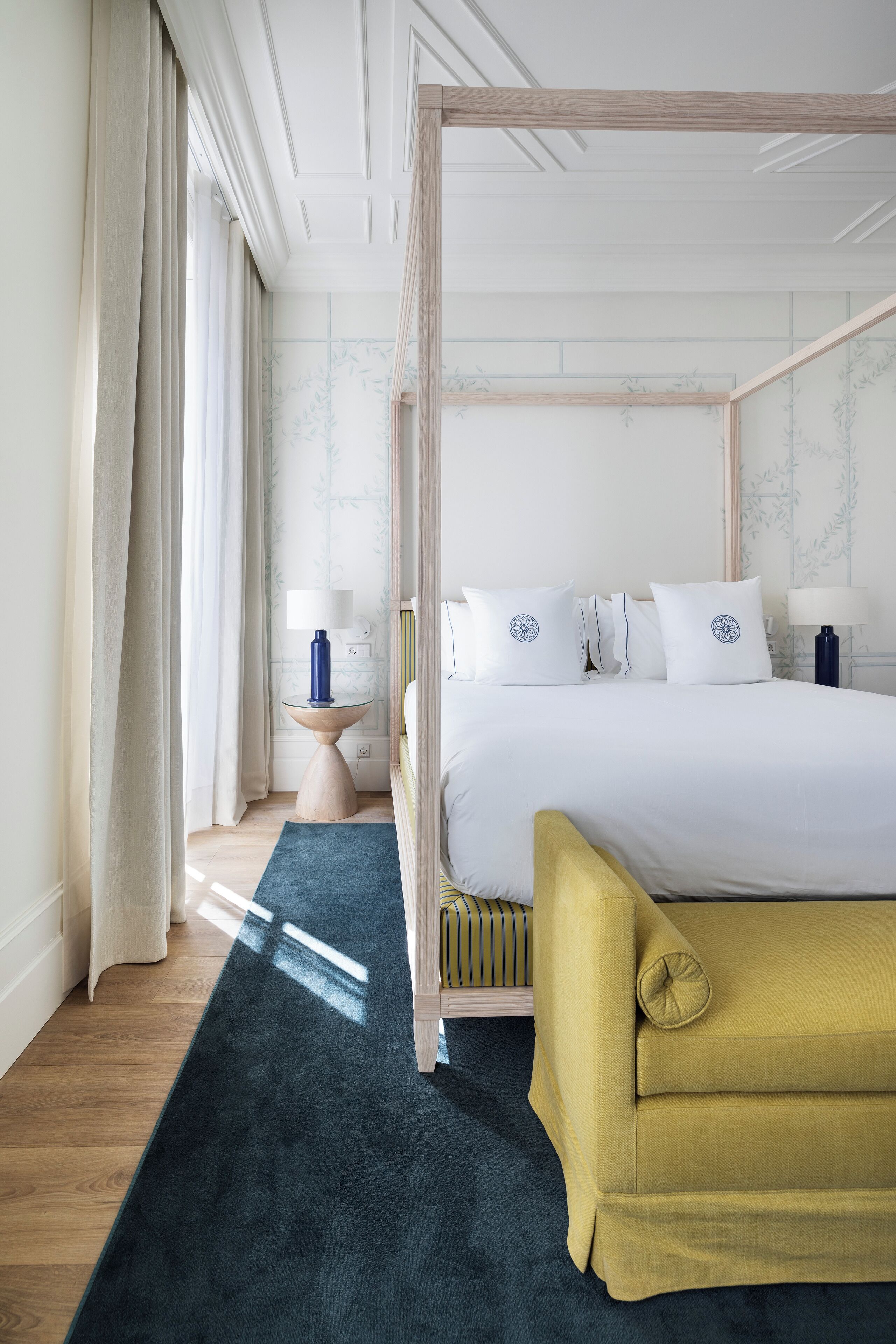 Een hemelbed met witte lakens en decoratieve kussens, een nachtkastje met een lamp, en een donkerblauwe kamerkleed.