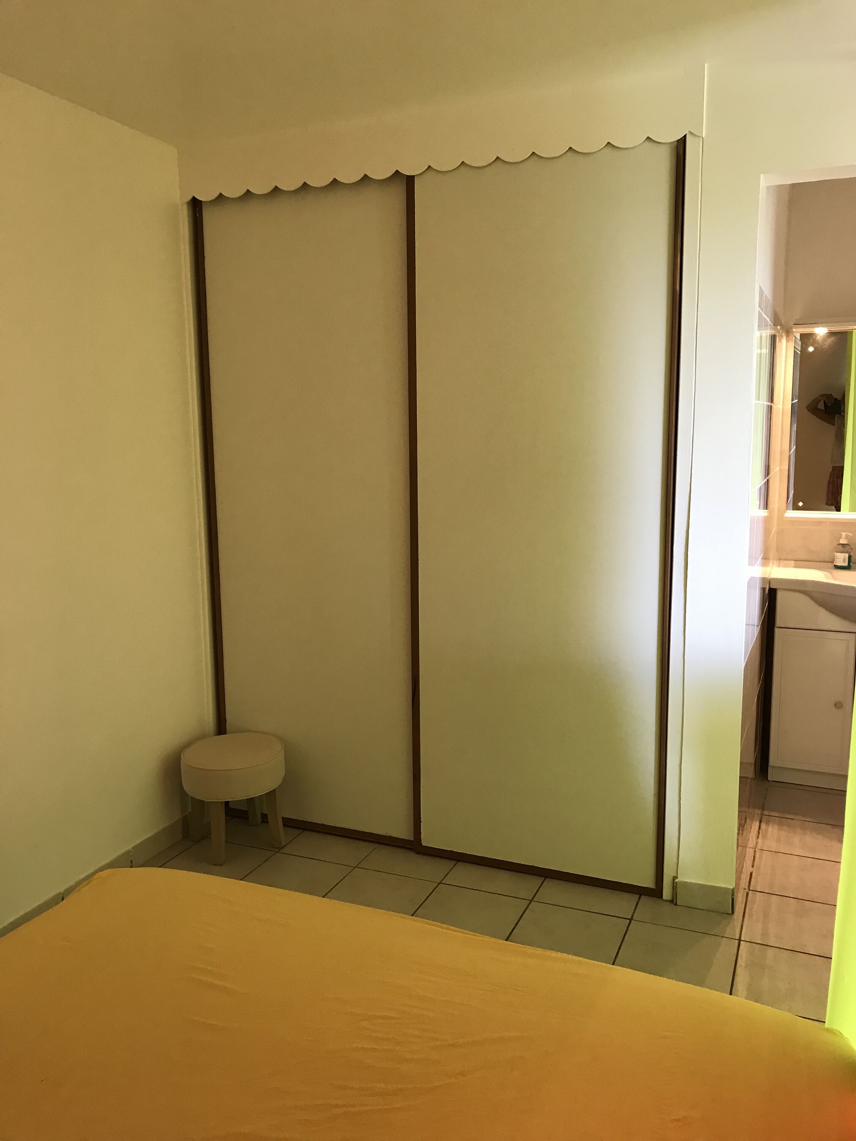 A bedroom with a bed, a small stool, a wardrobe, and a bathroom visible through a glass door.