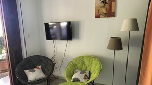 Een woonkamer met een zwarte bank, een groene fauteuil en een kleine groene tafel. Een televisie is aan de muur bevestigd, en er staat een vloerlamp.