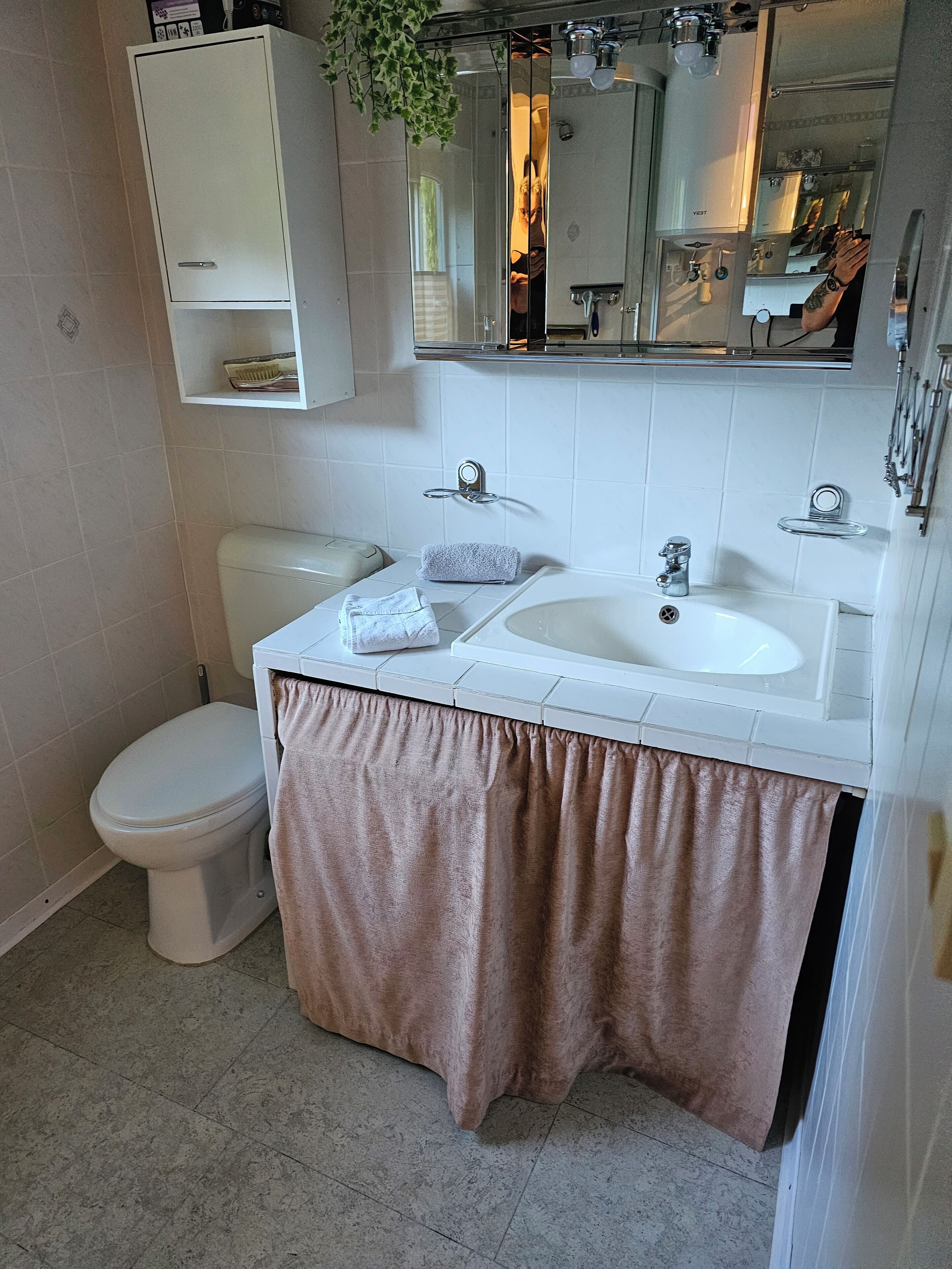 A bathroom with a toilet, sink, and mirror. There is a towel rack above the sink and a cabinet above the toilet.