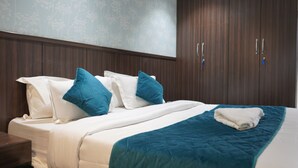 A neatly made bed with white and teal bedding, a folded white towel, and a wooden wardrobe with two doors. - Qotel Hotel Rajouri Garden (New Delhi)