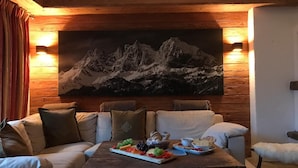 Una habitación acogedora con techo de madera, un cuadro grande de montañas, una mesa de madera con aperitivos y un sofá con cojines.