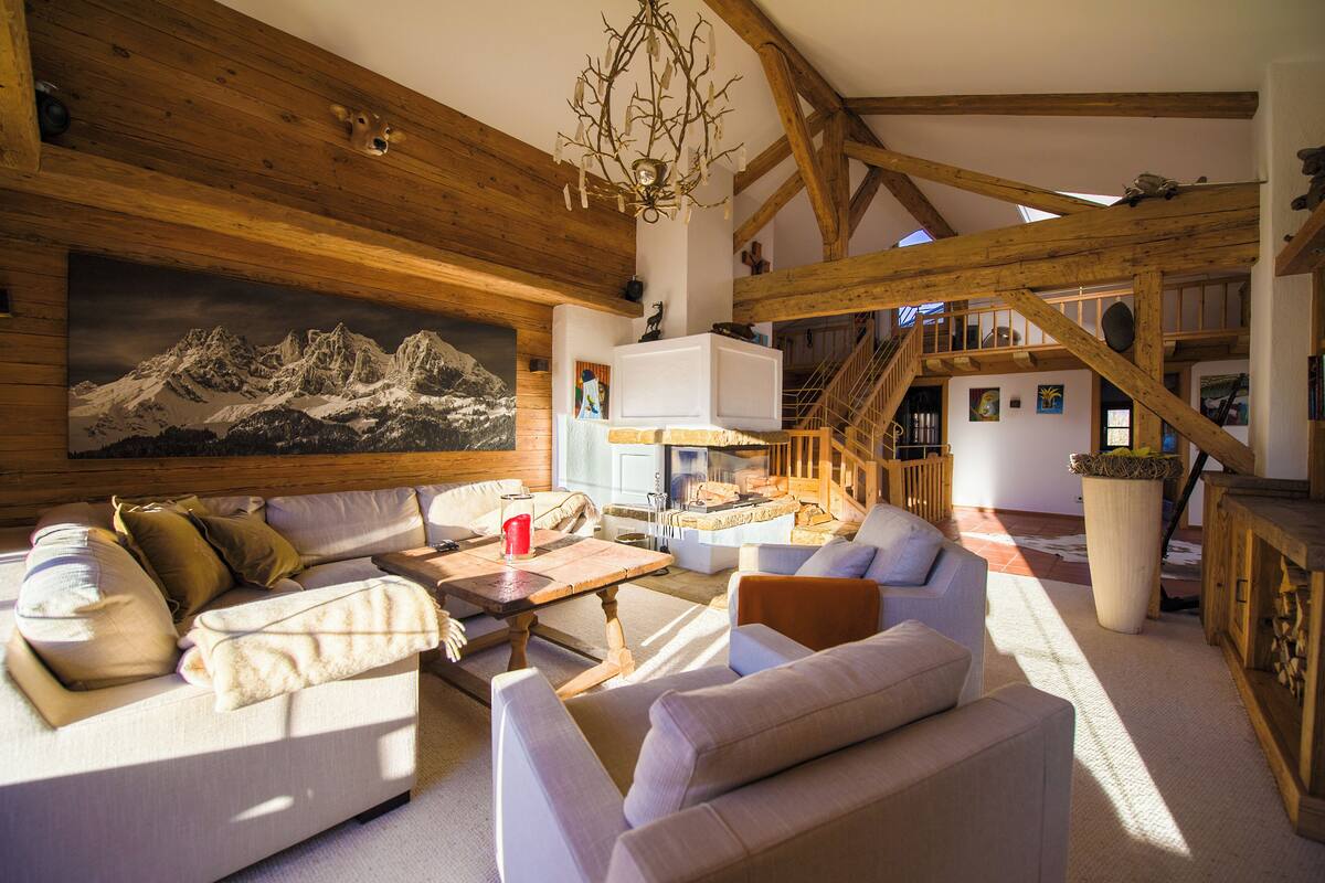 A spacious living area with a large painting of mountains, wooden beams, a staircase, and a cozy seating arrangement.