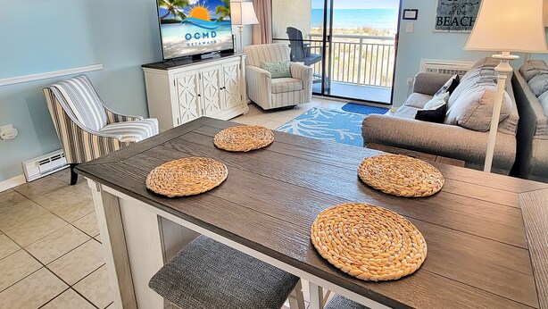 Een woonkamer met een houten eettafel, geweven placemats, een televisie en uitzicht op het strand door een schuifdeur.