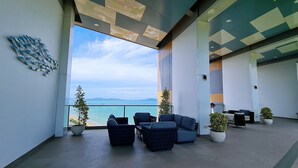 A modern lobby with a large glass wall offering a view of the sea and mountains. There are several dark-colored sofas and chairs arranged for seating. - Pattaya Seaview Beachfront Skypool Copacabana Jomtien Condo (Pattaya City)
