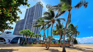A beach with palm trees and a modern building in the background. - Pattaya Seaview Beachfront Skypool Copacabana Jomtien Condo (Pattaya City)