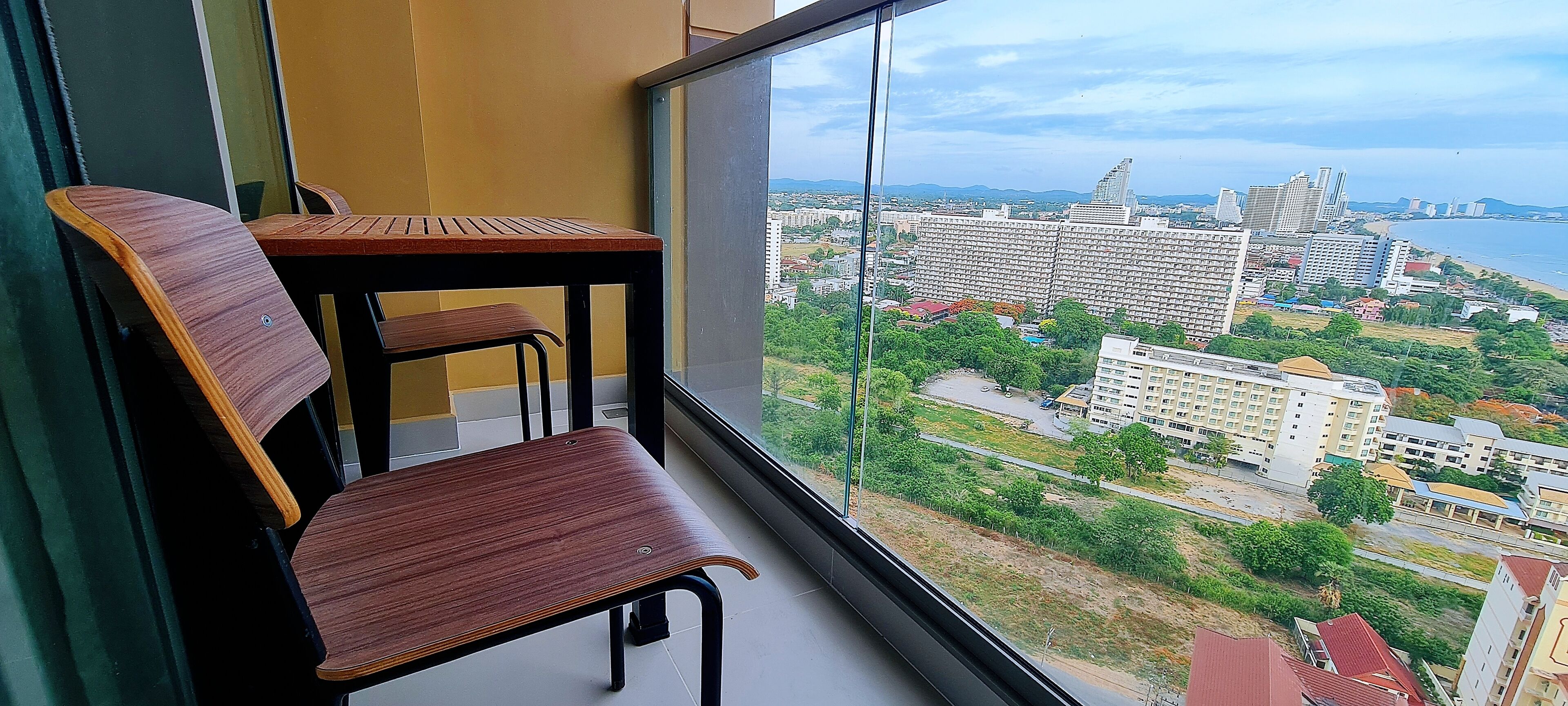 Un balcon avec deux chaises en bois et une table, d’où l’on peut admirer un paysage urbain avec des bâtiments et une étendue d’eau.