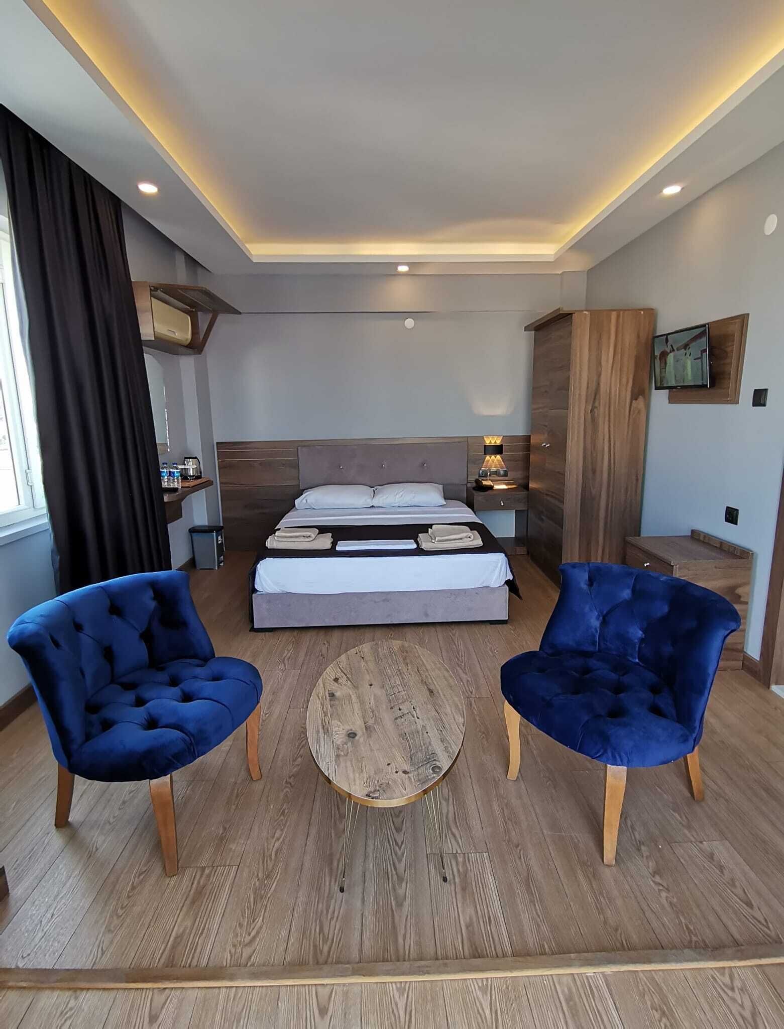 A modern hotel room with a bed, two blue armchairs, a round coffee table, and a TV mounted on the wall.