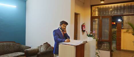 A man in a blue suit writing at a desk in a room with a blue wall, a wooden sofa, and a fire extinguisher on the wall.